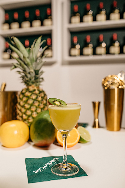 The kiwi canteen cocktail in a glass with a kiwi garnish sitting in front of a pineapple, orange, lime and other fruits