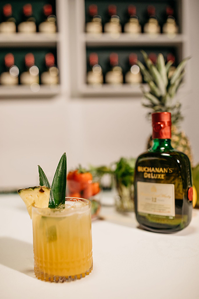 The piña crest drink garnished with a pineapple wedge in front of a Buchanan's Deluxe bottle 