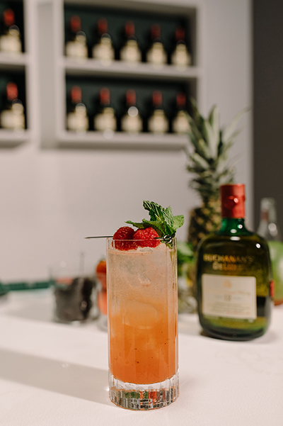 The signature raspberry cocktail garnished with 2 raspberries and a mint sprig in front of a bottle of Buchanan's Deluxe