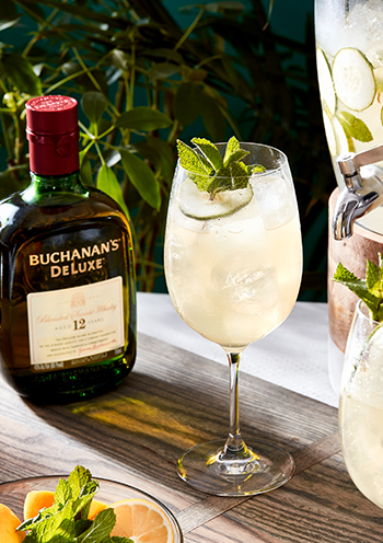 The summer is here cocktail garnished with a mint sprig and a cucumber in a wine glass in front of a bottle of Buchanan's Deluxe